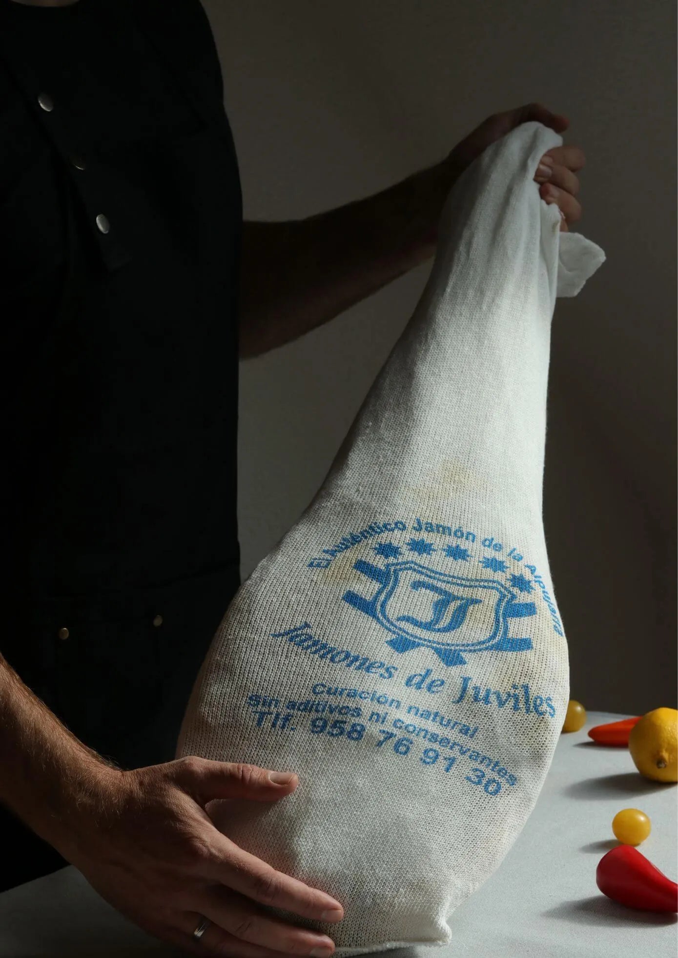 Ganze Schinkenkeule von Jamones de Juviles, traditionell verpackt im weißen Stoffbeutel mit blauer Aufschrift, in den Händen eines Verkäufers präsentiert.