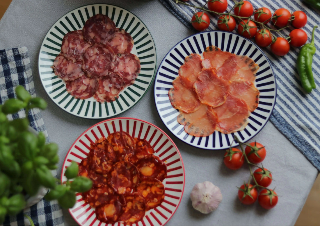Drei Teller mit spanischen Aufschnittsorten (Serrano, Chorizo, Lomo) auf einem gedeckten Tisch, umgeben von frischen Tomaten, Knoblauch und Kräutern.