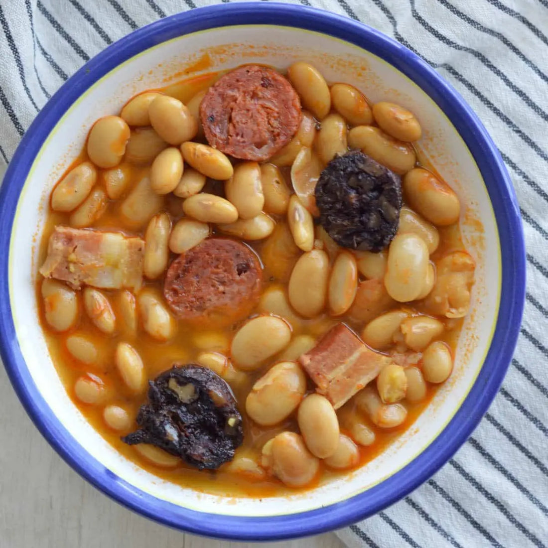 Traditionelle Fabada Asturiana mit weißen Bohnen in kräftiger Paprikabrühe, dazu Chorizo, Morcilla und Speckstücke in einer rustikalen Schüssel serviert.