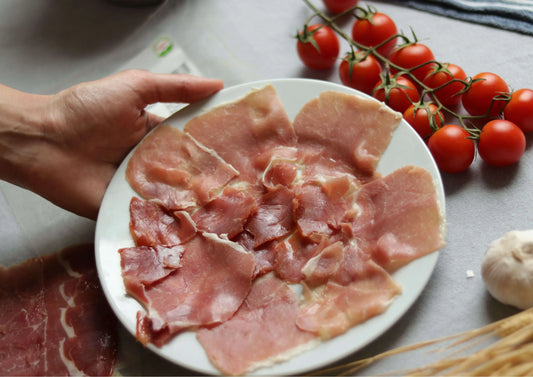 Teller mit dünn geschnittenem Serrano Schinken, gehalten von einer Hand, im Hintergrund frische Tomaten und Knoblauch.