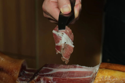 Hand hält mit einer Zange eine frisch geschnittene Scheibe spanischen Schinkens über einem Schinkenhalter.