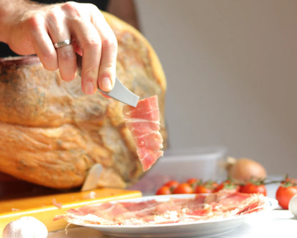 100 % Bio Serrano Jamón am Knochen, mit einem Schinkenmesser dünn aufgeschnitten, zart rosa Fleisch mit feiner Fettmarmorierung, serviert auf einem Teller mit Tomaten im Hintergrund.
