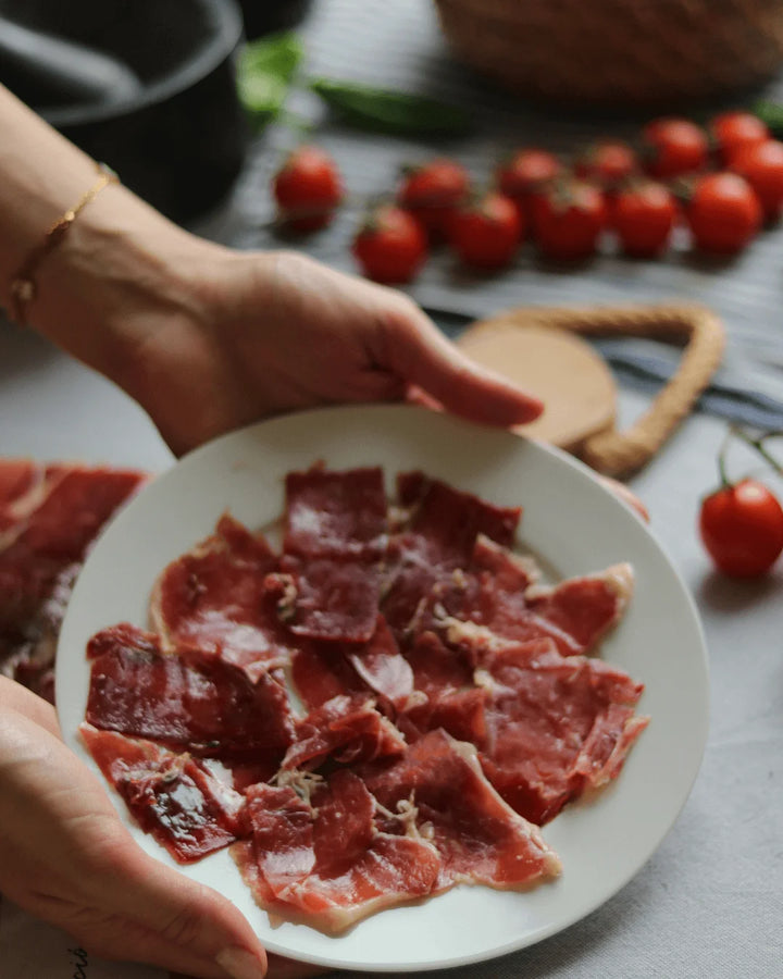 Aufgeschnittene Ibérico-Schinkenscheiben auf einem weißen Teller, von zwei Händen gehalten, im Hintergrund Tomaten und Küchenutensilien.