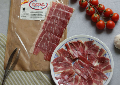 Aufgeschnittener Jamón Serrano Gran Reserva (Schinken) angerichtet auf einem Teller, daneben Packung mit Etikett, Kirschtomaten und Knoblauch