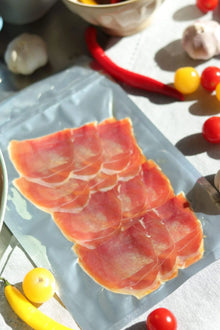 Aufschnittpackung mit dünn geschnittenem Serrano Schinken, umgeben von frischem Gemüse und Chili.