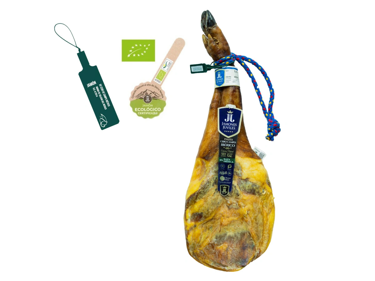 Produktbild einer oekologischen Bio Paleta de Cebo de Campo Iberica von Jamones Juviles vor weissem Hintergrund. Die iberische Vorderschulter verfügt ueber eine dunkle Klaue, eine blau-gelbe Kordel und das blaue Markenetikett. Links im Bild sind die offizielle dunkelgruene Siegelplombe, das grüne EU-Bio-Logo und das Zertifikat fuer oekologische Erzeugung in der Sierra Nevada abgebildet.