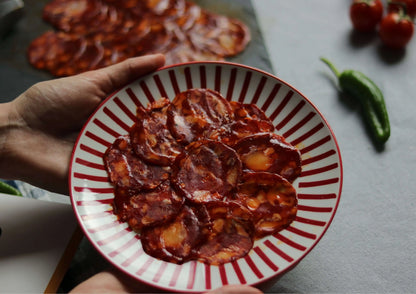 Chorizo Ibérico in Scheiben, angerichtet auf einem rot-weiß gestreiften Teller, von Händen gehalten, im Hintergrund weitere Chorizoscheiben, Tomaten und eine grüne Paprikaschote.