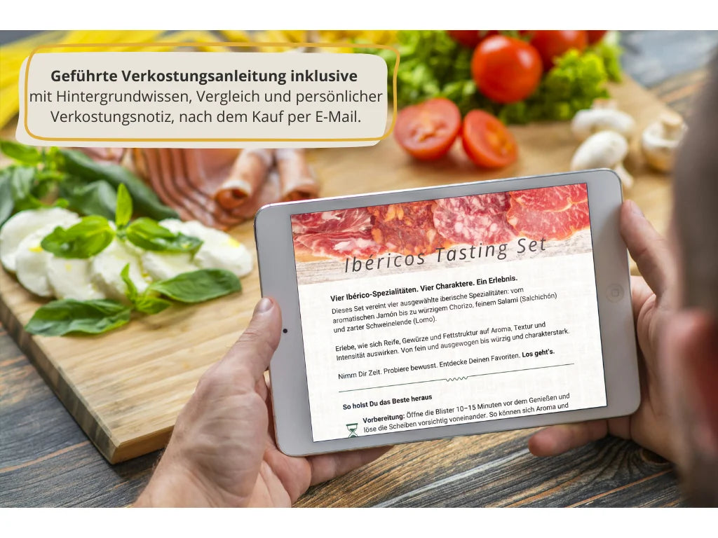 Tablet mit der digitalen Verkostungsanleitung des Ibéricos Tasting Sets, umgeben von spanischen Spezialitäten wie Chorizo, Salchichón, Jamón und frischen Zutaten auf einem Holztisch