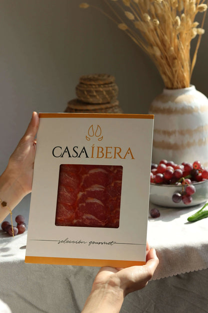 Eine Hand hält eine Packung „Casa Íbera“ mit dünn geschnittener Schweinelende (Lomo Ibérico). Die Scheiben sind gleichmäßig rot mit feiner Marmorierung sichtbar durch das Sichtfenster.