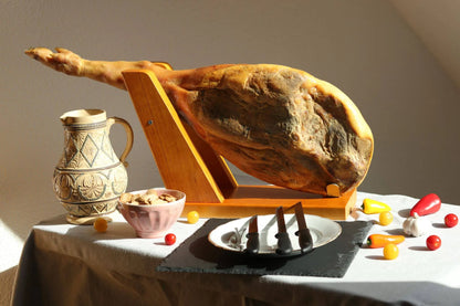 Ganze spanische Schinkenkeule (Jamón) in einem Holzschinkenhalter, servierfertig mit Messern, traditionellem Krug und Beilagen wie Knoblauch, Paprika und Kirschtomaten auf einem gedeckten Tisch. 