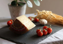 Gereifter Mischkäse (Queso de Mezcla Curado) auf Schieferplatte mit brauner Rinde, dazu rote Cherrytomaten und mediterrane Dekoration im Hintergrund