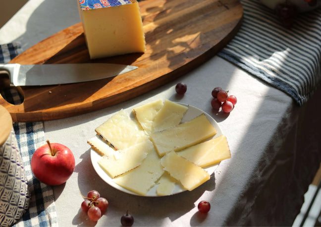 Gereifter Schafskäse (Queso Curado de Oveja) in Scheiben auf einem Teller, daneben ein größeres Stück Käse auf einem Holzbrett mit Messer, dekoriert mit Trauben und einem Apfel.