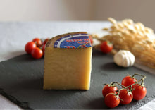 Gereifter Schafskäse (Queso de Oveja Curado) auf Schieferplatte mit Tomaten, Knoblauch und Ähren im Hintergrund.