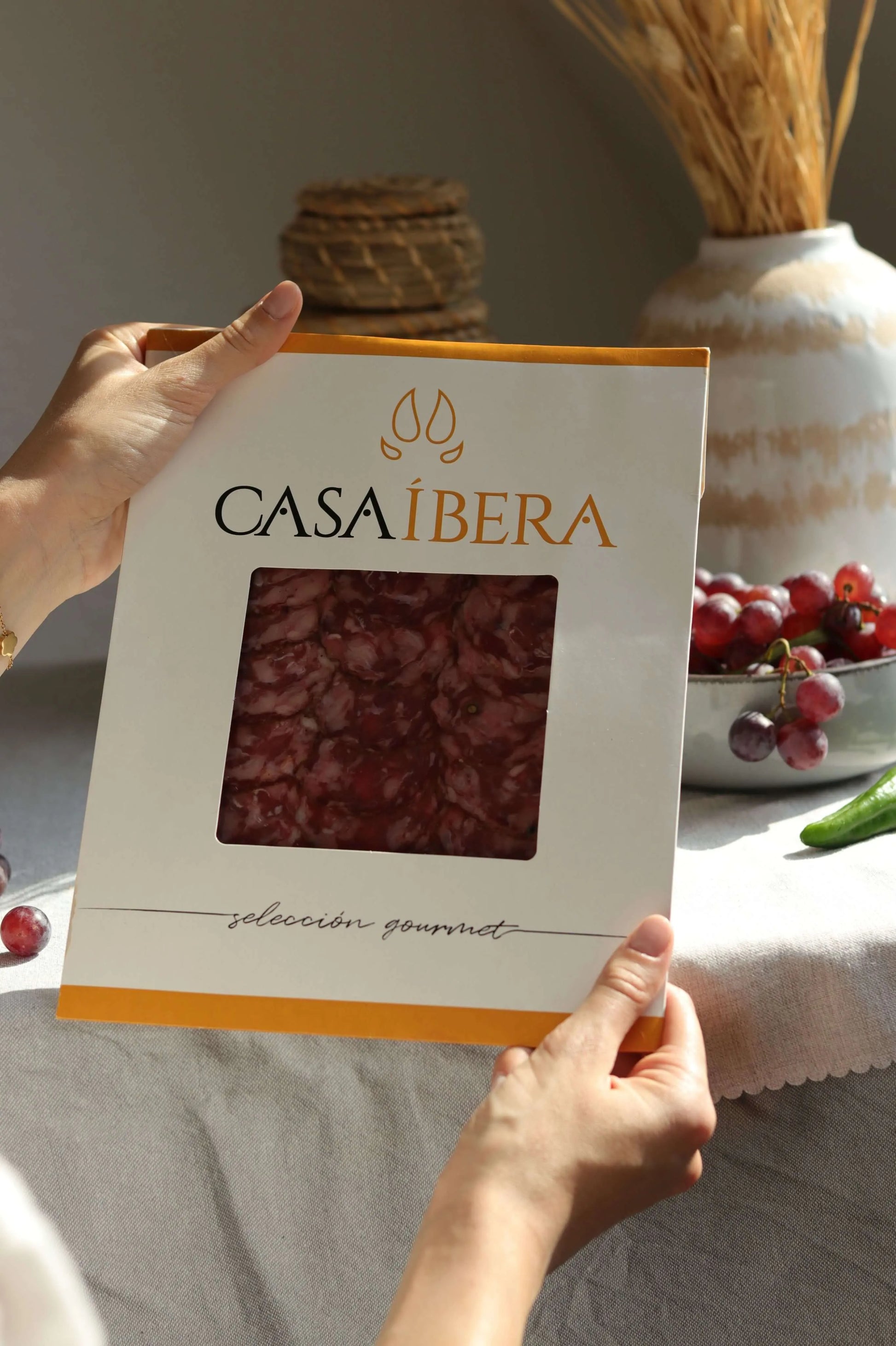 Hände halten eine Packung Casa Íbera mit aufgeschnittenem Ibérico Salami (Salchichón), Gourmet-Auswahl, auf einem Tisch mit Trauben und Deko im Hintergrund.
