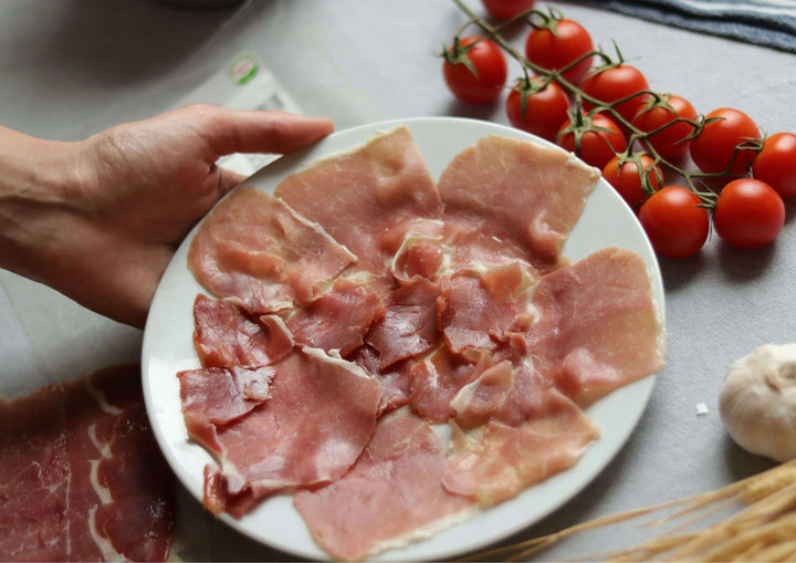 Hand hält einen Teller mit Serrano-Schinken Reserva (Jamón Serrano), im Hintergrund Strauchtomaten und Knoblauch.