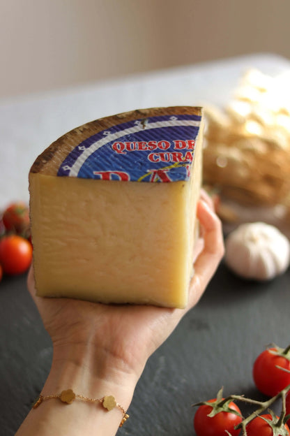 Hand hält ein Stück gereiften Schafskäse (Queso de Oveja Curado) mit blauem Etikett, dekoriert mit frischen Tomaten.