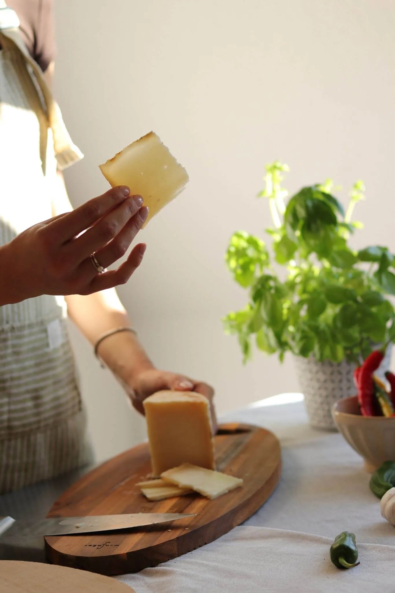 Hand hält eine Scheibe spanischen Schafskäses, im Hintergrund ein Holzbrett mit dem restlichen Käse, Basilikum-Pflanze und bunte Chilis – mediterrane Atmosphäre.