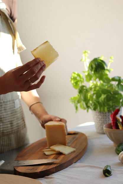 Hand hält eine Scheibe spanischen Schafskäses, im Hintergrund ein Holzbrett mit dem restlichen Käse, Basilikum-Pflanze und bunte Chilis – mediterrane Atmosphäre.