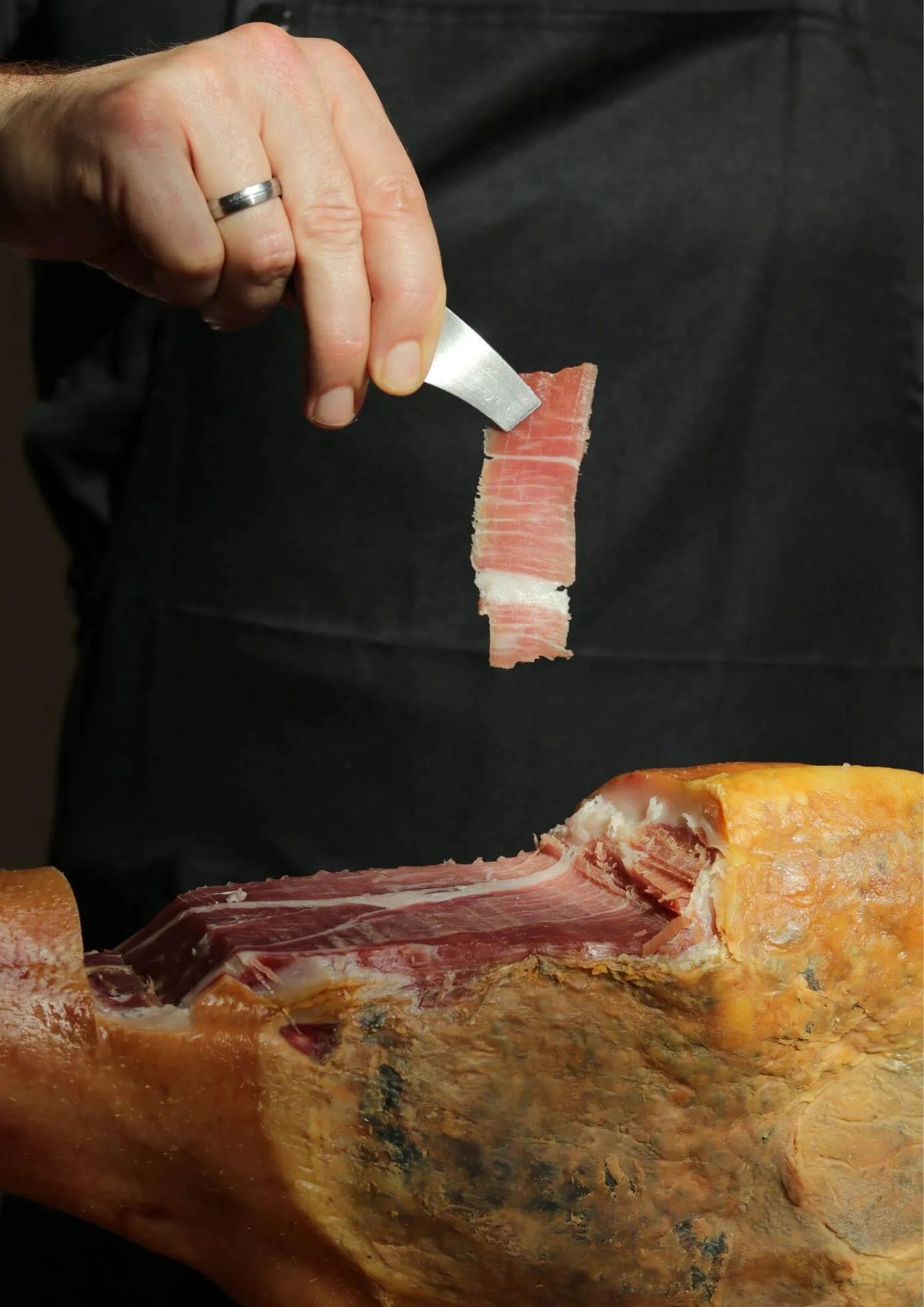 Hand hält mit einer Pinzette eine feine Scheibe spanischen Schinkens über einem aufgeschnittenen Schinkenstück im Schinkenhalter