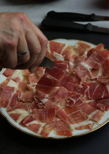 Hand serviert eine Scheibe fein geschnittenen Schinkens auf einem Teller mit vielen Jamón-Scheiben.