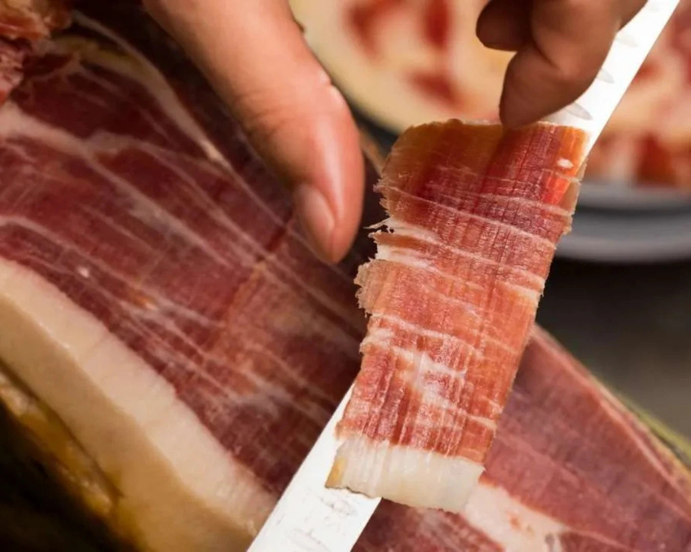 Ibérico Cebo Jamón als dünne Scheibe auf einem Schinkenmesser, rot schimmerndes Fleisch mit gleichmäßiger Fettmarmorierung, im Hintergrund ein angeschnittener Ibérico-Schinken.