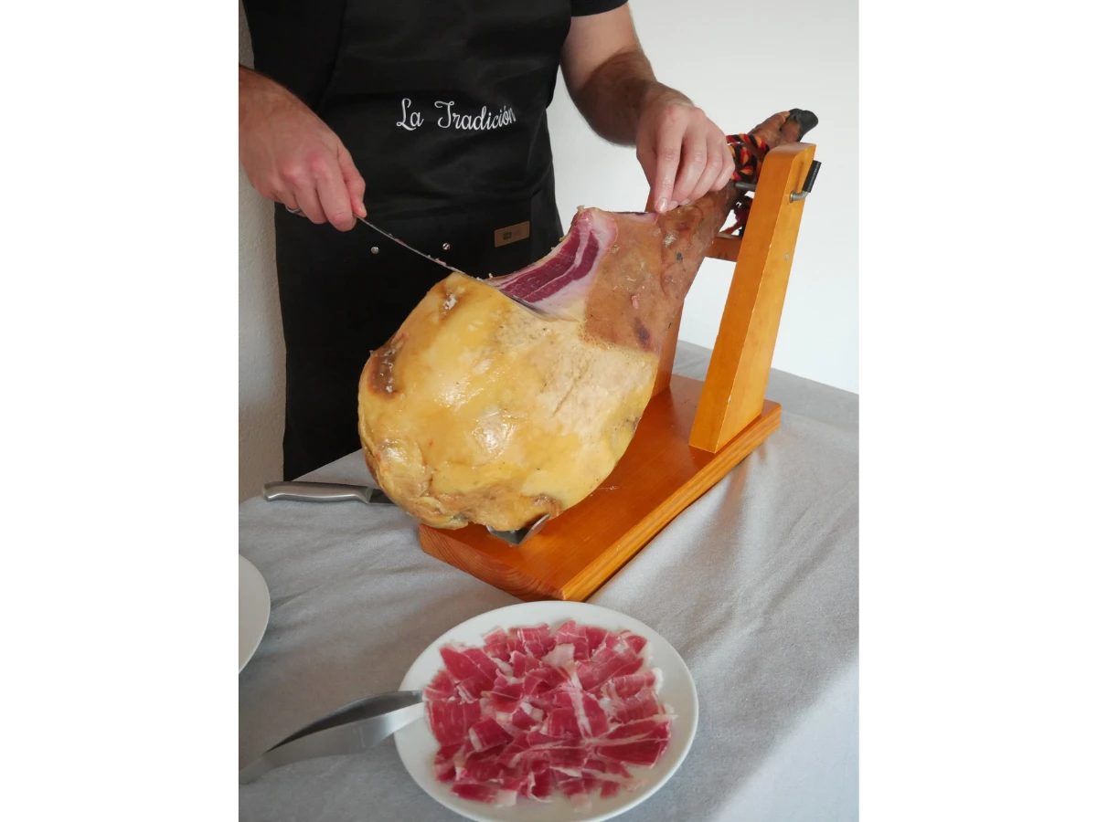 Person schneidet Jamón Ibérico Pata Negra auf einem Holz-Schinkenhalter, darunter ein Teller mit frisch geschnittenen Scheiben.