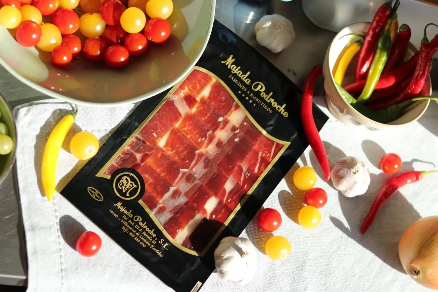 Jamón Ibérico Pata Negra D.O.P. Los Pedroches, handgeschnittene Aufschnittpackung von Majada Pedroche, dekoriert mit Tomaten, Knoblauch und Chilischoten auf weißem Tuch.