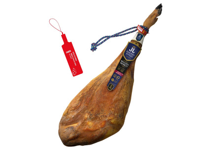 Produktbild eines ganzen Jamon de Bellota Iberico der Premiummarke Jamones Juviles auf weissem Grund. Der edle Schinken praesentiert sich in gestreckter Form mit dunkler Klaue, einer blau-roten Kordel und einem eleganten dunkelblauen Etikett. Links daneben ist die offizielle rote Siegelplombe zu sehen, die gesetzliche Kennzeichnung fuer spanischen Iberico Bellota Schinken aus traditioneller Eichelmast.