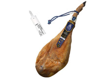 Produktbild eines ganzen Jamon de Cebo Iberico von Jamones Juviles auf weissem Grund. Der luftgetrocknete Hinterschinken praesentiert sich in einer eleganten Form mit blau-roter Kordel und dem dunkelblauen Markenetikett der Manufaktur. Links im Bild wird die offizielle weisse Siegelplombe dargestellt, die gesetzliche Kennzeichnung fuer garantierten spanischen Iberico Schinken aus Cebo-Haltung.