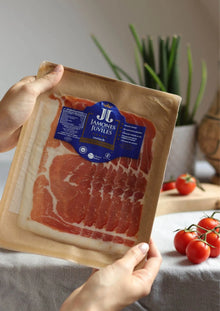 Jamón de Trevélez D.O.P. Serrano Schinken in Scheiben 100 g Packung, gehalten in der Hand mit frischen Tomaten im Hintergrund