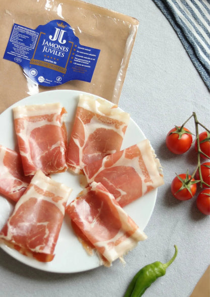 Jamón de Trevélez D.O.P. Serrano Schinken in Scheiben, serviert auf Teller mit Tomaten im Hintergrund
