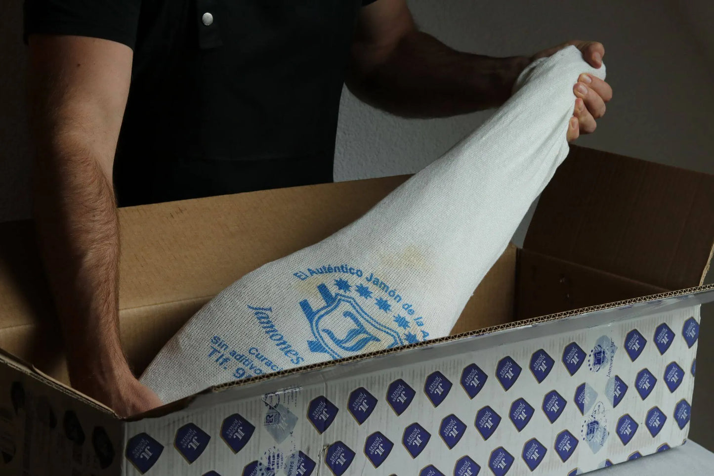 Mann packt einen ganzen Schinken von Jamones de la Alpujarra in einer weißen Stoffhülle aus einem Karton mit Markenlogo aus.