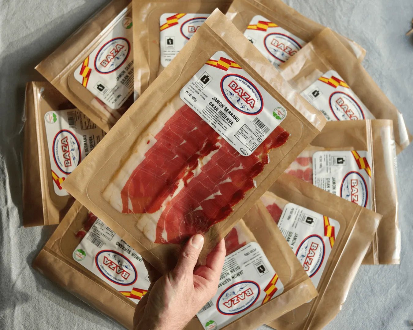 Mehrere Packungen spanischer Jamón Serrano Gran Reserva der Marke Baza, nachhaltige Papierverpackung, eine Packung wird in der Hand gehalten; hochwertiger luftgetrockneter Serranoschinken aus Spanien.