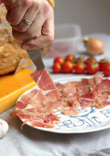 Nahaufnahme einer Hand mit Ring, die eine Pinzette hält und eine dünne Scheibe spanischen Schinkens (Jamón) von einem Teller mit mehreren Schinkenscheiben aufnimmt. Im Hintergrund sind Tomaten zu sehen