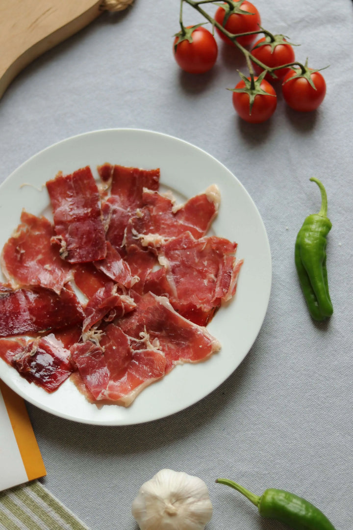 Nahaufnahme von Jamón Ibérico (Schinken) Scheiben auf weißem Teller, serviert mit Gemüse-Dekoration auf einer Tischdecke.