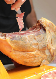 Nahaufnahme einer ganzen Schinkenkeule in einem Holz-Jamonero, während eine Hand mit einer Pinzette mehrere frisch geschnittene, dünne Scheiben spanischen Schinkens (Jamón) hochhält.