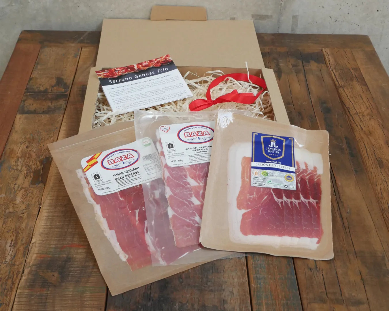 Offene Geschenkbox „Serrano Genuss Trio“ mit drei vakuumverpackten spanischen Serrano-Schinken (Baza und Trevélez), dekoriert mit naturfarbenem Füllmaterial und roter Schleife. Spanisches Feinkost-Set als Geschenkidee