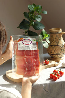 Packung Jamón Serrano (Schinken) Baza, dünn aufgeschnitten und vakuumverpackt. In den Händen gehalten, mit Keramikkrug, Tomaten und Knoblauch im Hintergrund auf einem gedeckten Tisch.