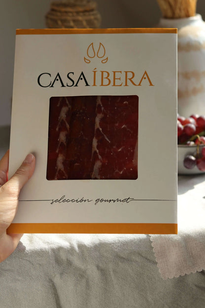Packung „Casa Íbera“ mit feinen Scheiben von Jamón Ibérico (Schinken). Der Aufschnitt zeigt eine intensive rote Farbe mit weißer Fettmarmorierung, erkennbar durch das Sichtfenster.