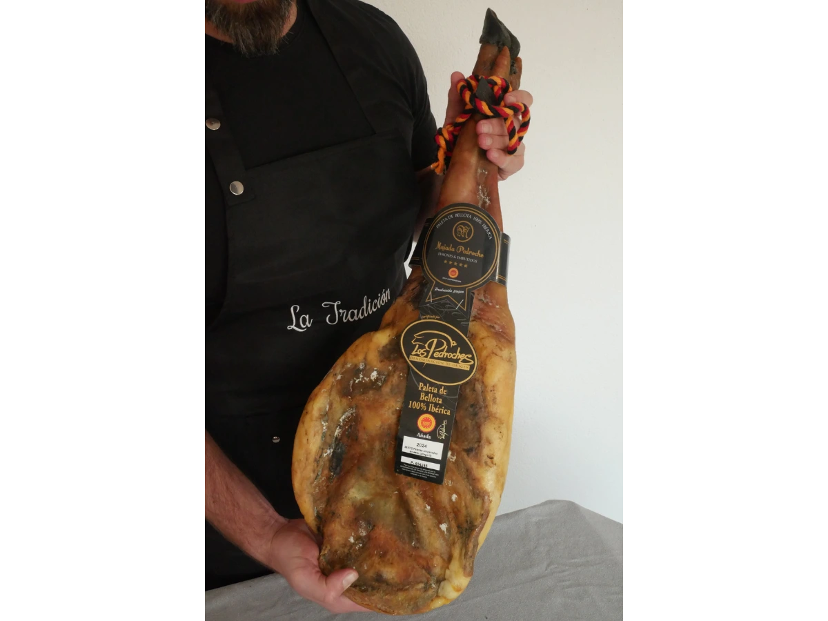 Person mit Schürze „La Tradición“ hält eine ganze Paleta Pata Negra 100 % Ibérico von Los Pedroches mit schwarzer Bellota-Etikette.