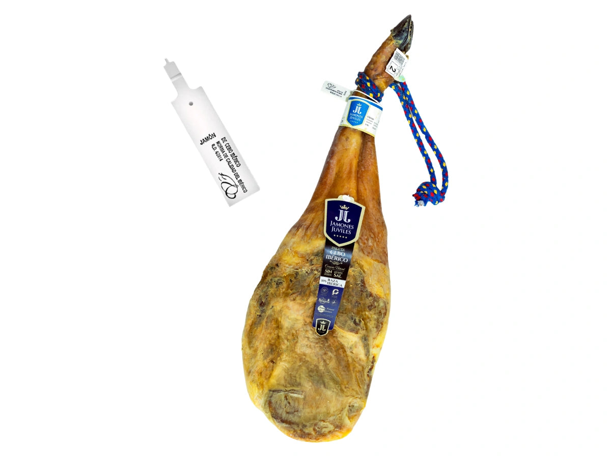 Produktbild einer ganzen Paleta de Cebo Iberica von Jamones Juviles vor weissem Hintergrund. Die iberische Vorderschulter zeigt eine naturbelassene Form mit blau-gelber Kordel am Hufeisen und einem dunkelblauen Markenetikett auf dem Stueck. Links daneben ist die offizielle weisse Kunststoffplombe abgebildet, die die gesetzliche Qualitaetskennzeichnung fuer De Cebo Iberico nach der spanischen Norm garantiert.