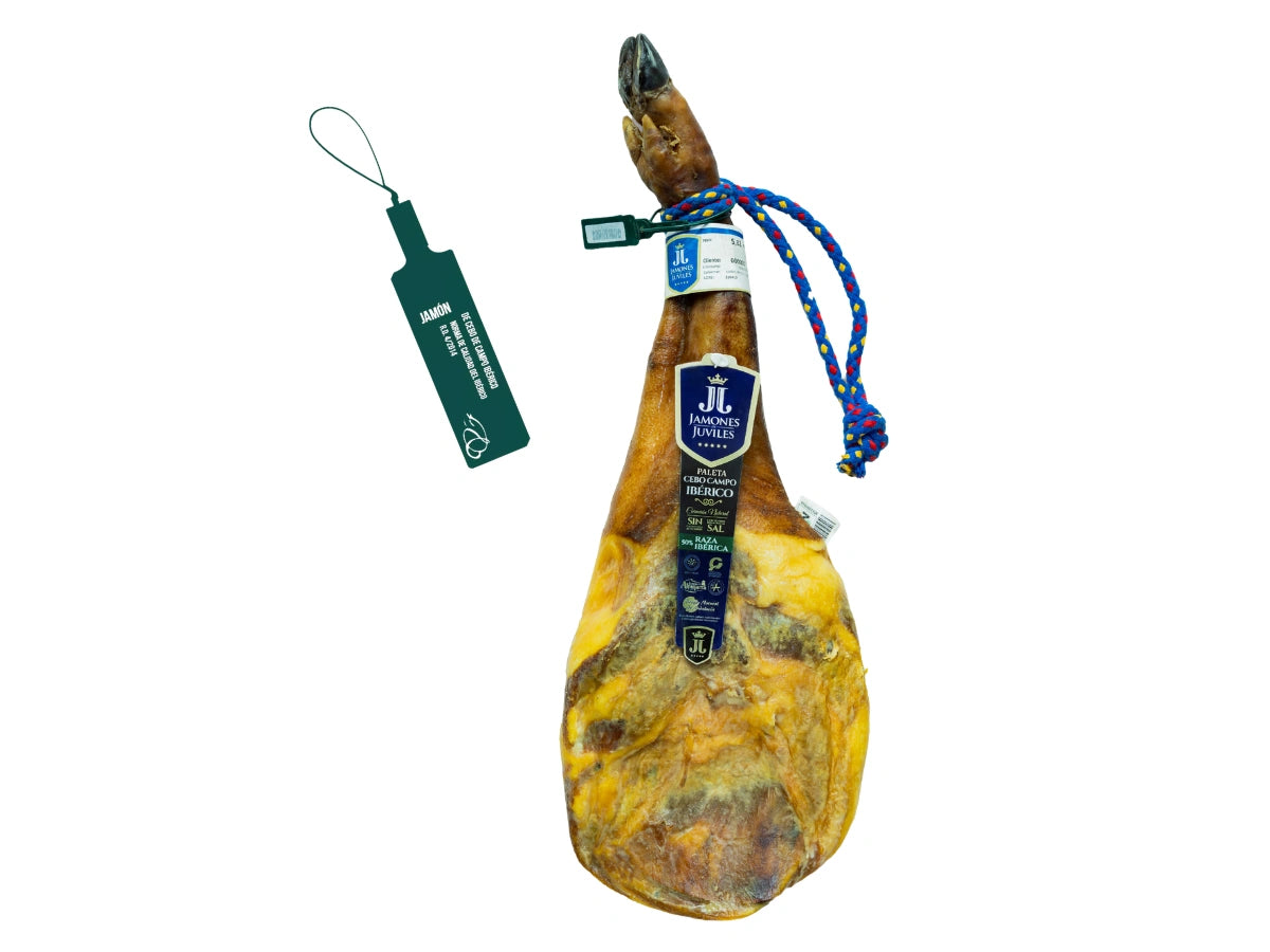 Produktbild einer ganzen Paleta de Cebo de Campo Iberica von Jamones Juviles vor weissem Hintergrund. Die iberische Vorderschulter hat eine dunkle Klaue, eine blau-gelbe Kordel und ein dunkelblaues Markenetikett auf dem Stueck. Links daneben wird die offizielle dunkelgruene Kunststoffplombe abgebildet, welche die gesetzliche Qualitaetskennzeichnung fuer Cebo de Campo Iberico gemaess der spanischen Norm garantiert.