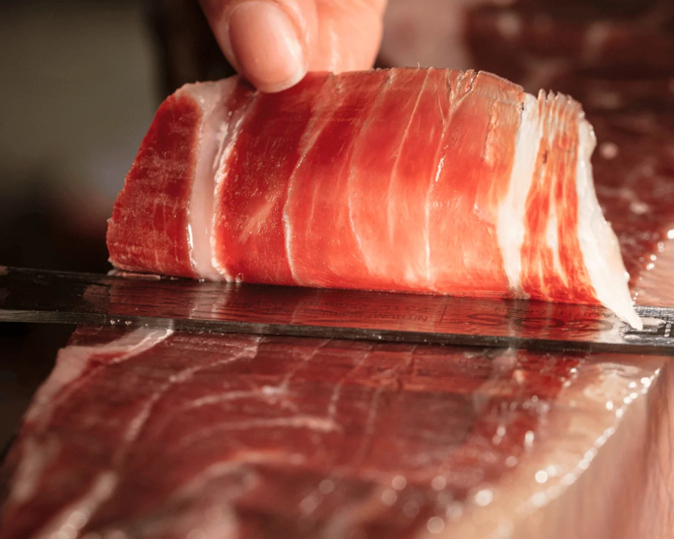 Pata Negra Ibérico Jamón in einer dünnen Scheibe auf einem Schinkenmesser, intensiv dunkelrote Fleischfarbe mit feiner Fettmarmorierung, glänzende Oberfläche durch lange Reifung.