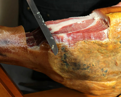 Serrano Reserva Jamón am Knochen, frisch mit einem langen Schinkenmesser angeschnitten, sichtbare Marmorierung und gereifte Kruste des luftgetrockneten spanischen Schinkens.