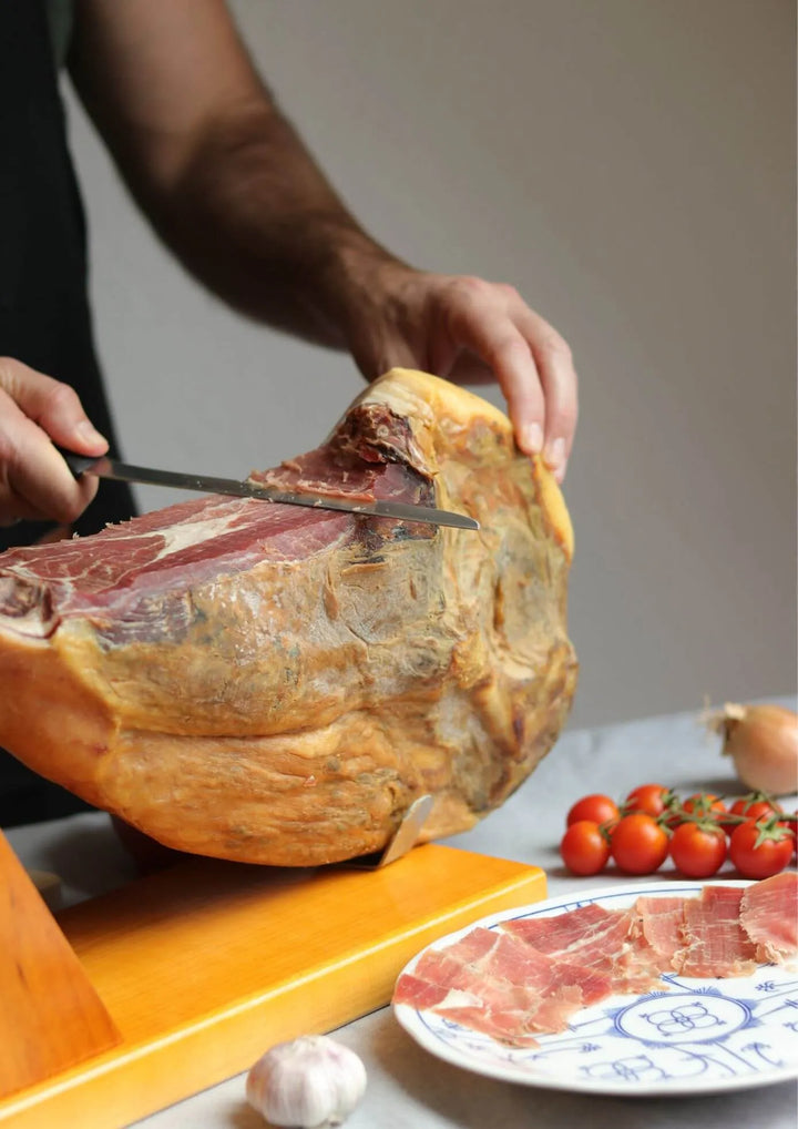 Spanische Schinkenkeule auf einem Holz-Jamonero, von Hand in dünne Scheiben geschnitten. Im Vordergrund ein Teller mit Jamón-Scheiben, daneben Knoblauch und frische Tomaten als Beilage. 