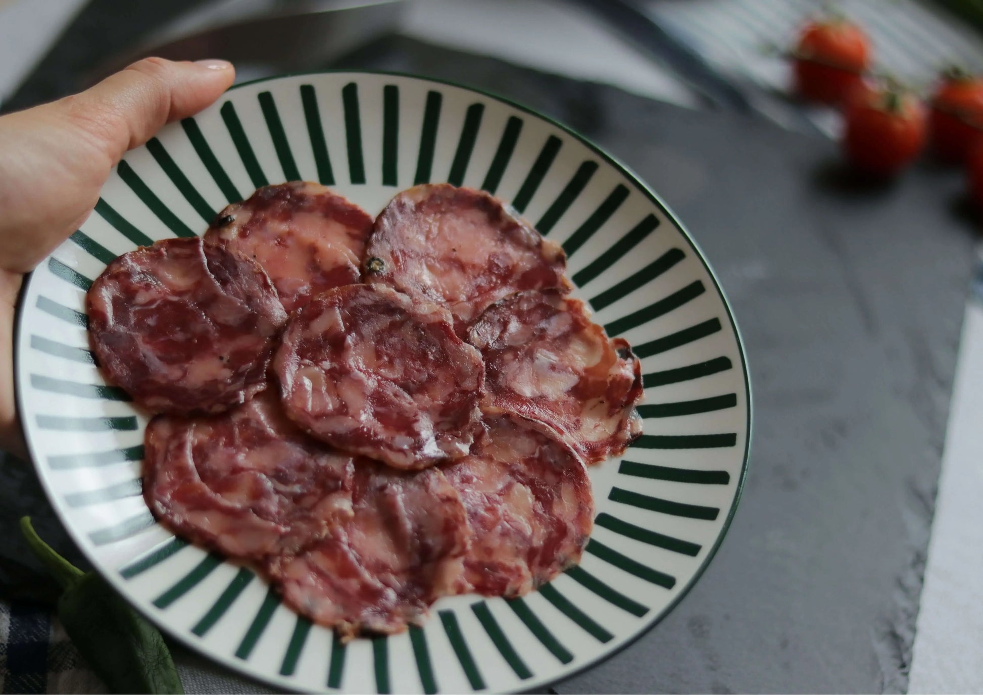 Chorizo Ibérico in Scheiben, angerichtet auf einem rot-weiß gestreiften Teller, von Händen gehalten, im Hintergrund weitere Chorizoscheiben, Tomaten und eine grüne Paprikaschote.