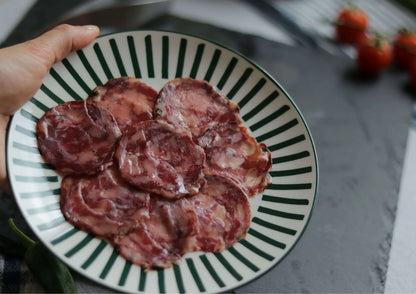 Chorizo Ibérico in Scheiben, angerichtet auf einem rot-weiß gestreiften Teller, von Händen gehalten, im Hintergrund weitere Chorizoscheiben, Tomaten und eine grüne Paprikaschote.