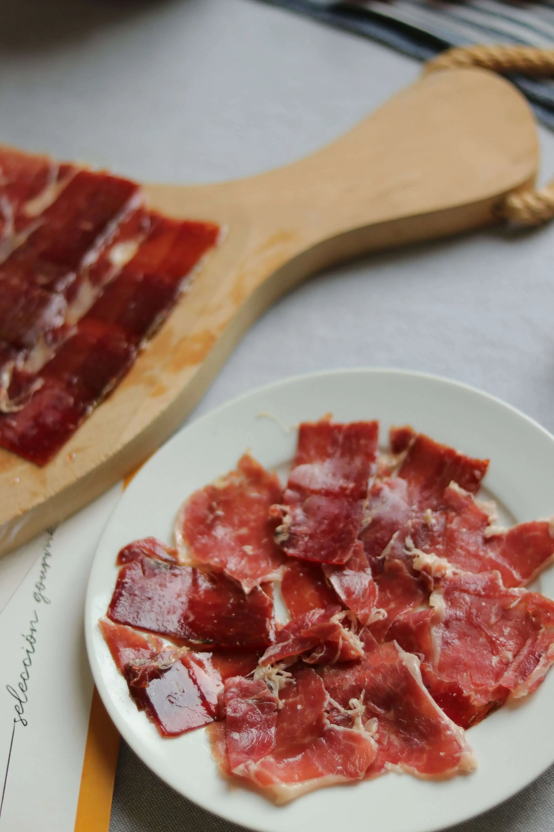Teller mit dünn geschnittenem Jamón Ibérico (Schinken), angerichtet auf weißem Teller mit Tomaten und grünem Paprika im Hintergrund.
