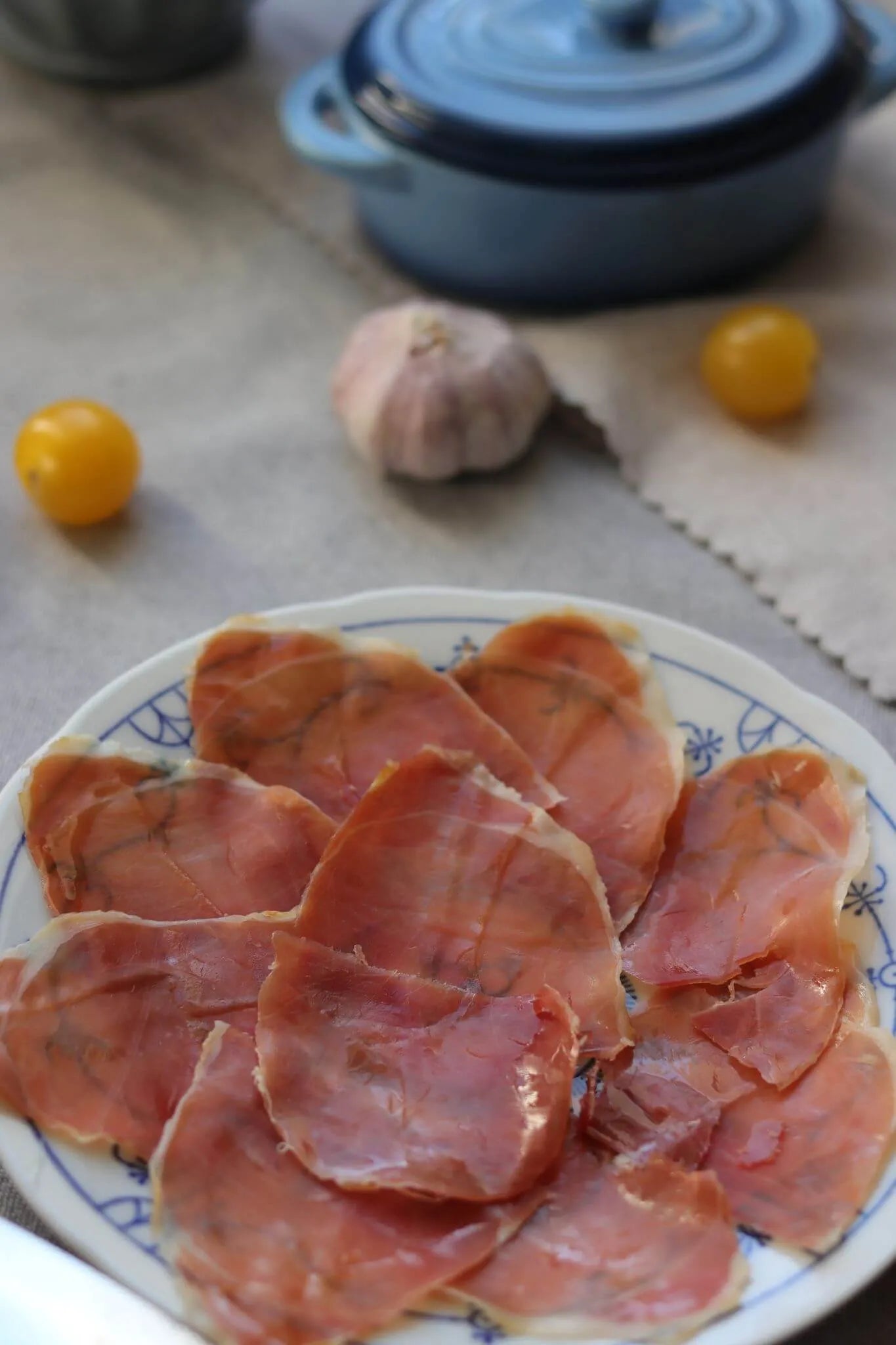 Teller mit dünn geschnittenem Serrano Schinken, angerichtet auf einem weißen Teller mit blauem Muster, im Hintergrund Knoblauch und gelbe Tomaten.