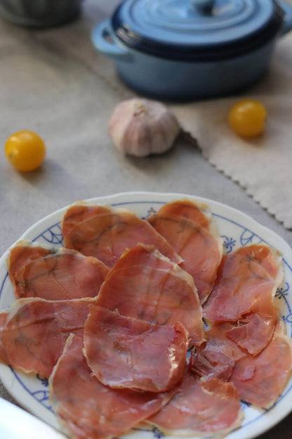 Teller mit dünn geschnittenem Serrano Schinken, angerichtet auf einem weißen Teller mit blauem Muster, im Hintergrund Knoblauch und gelbe Tomaten.